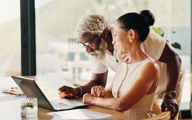 A couple in their sixties looks over a laptop and printed paperwork together at a table at home.